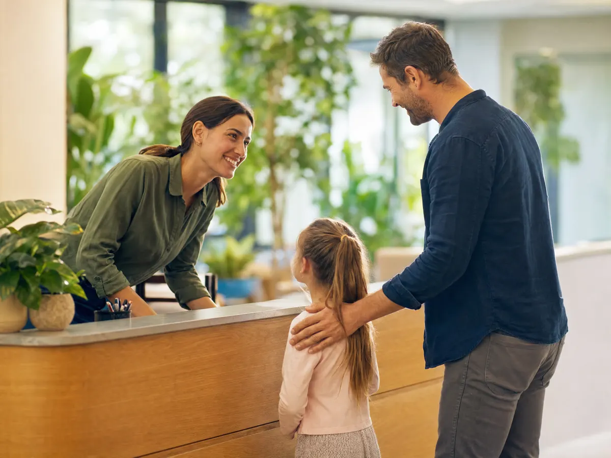 A front-desk staff member welcoming a father and his young daughter in a bright reception area